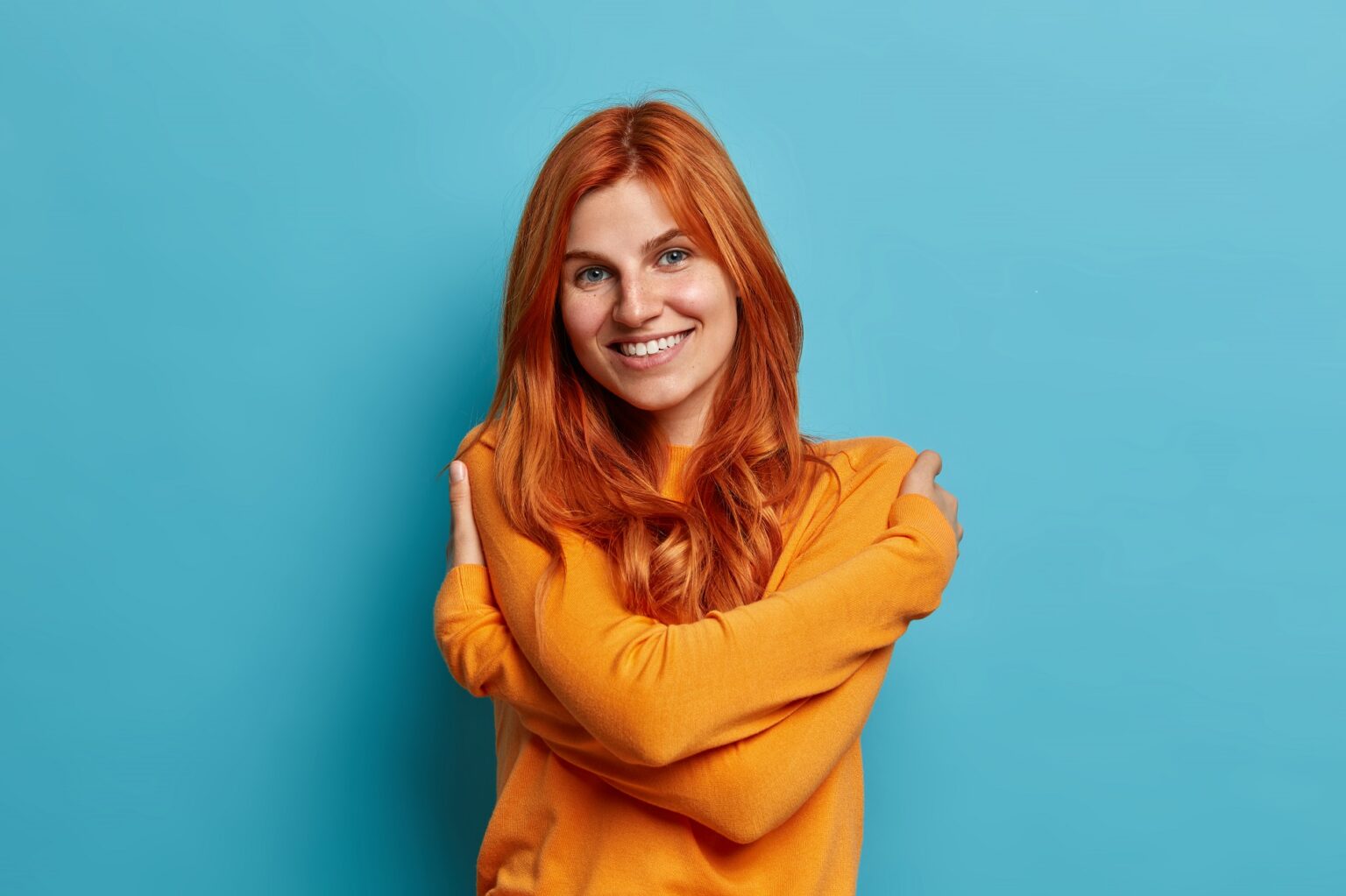 Charming pleased redhead european woman embraces own body dressed in casual orange jumper smiles pleasantly feels comfortable poses against blue background. tenderness and self esteem concept